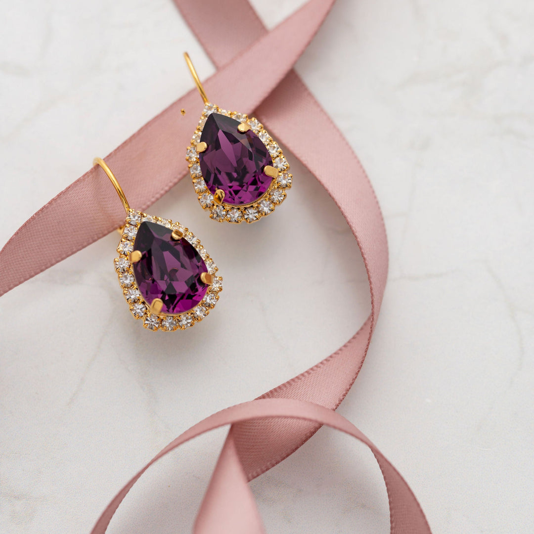 Pair of purple crystal earrings with gold accents on a pink ribbon against a white background